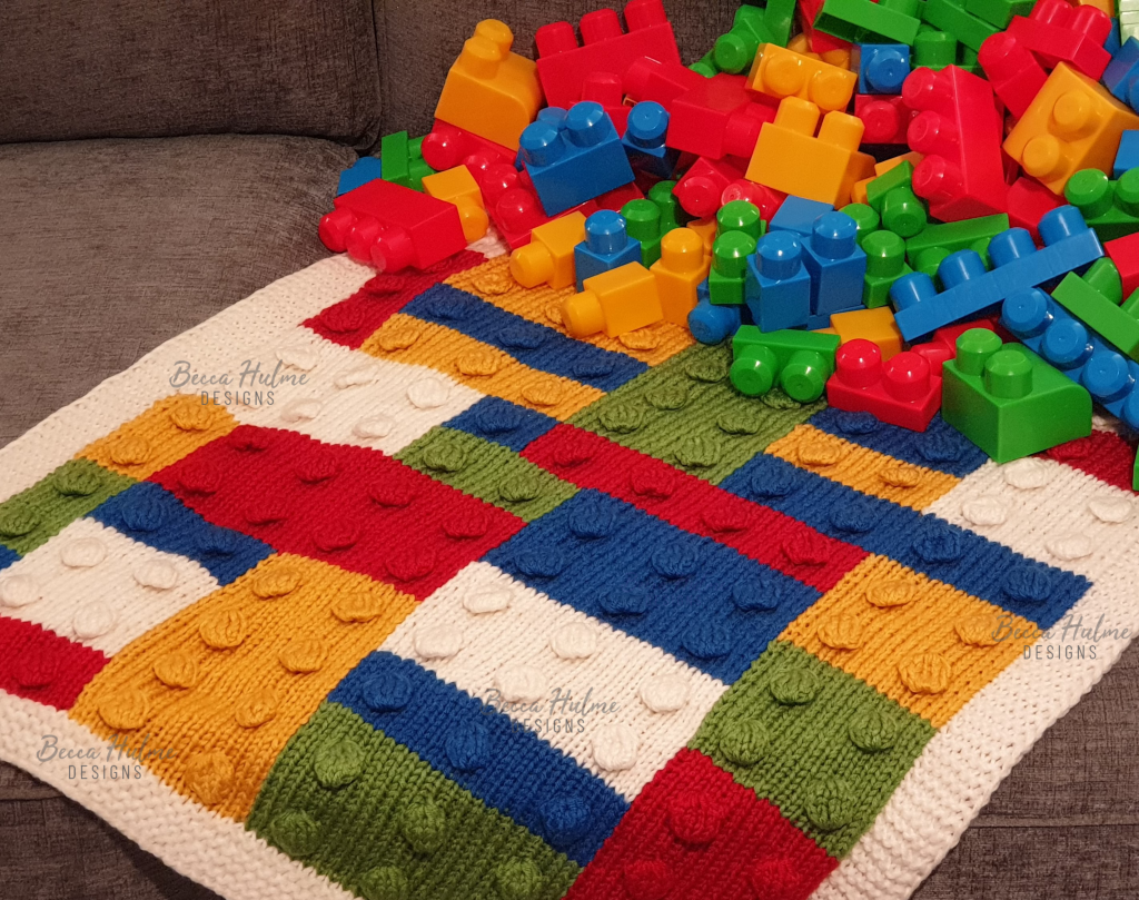 Colourful knitted blanket draped on a sofa with a pile of kids building blocks at one end. The blanket design mimics the blocks with bobbles and colour layout.