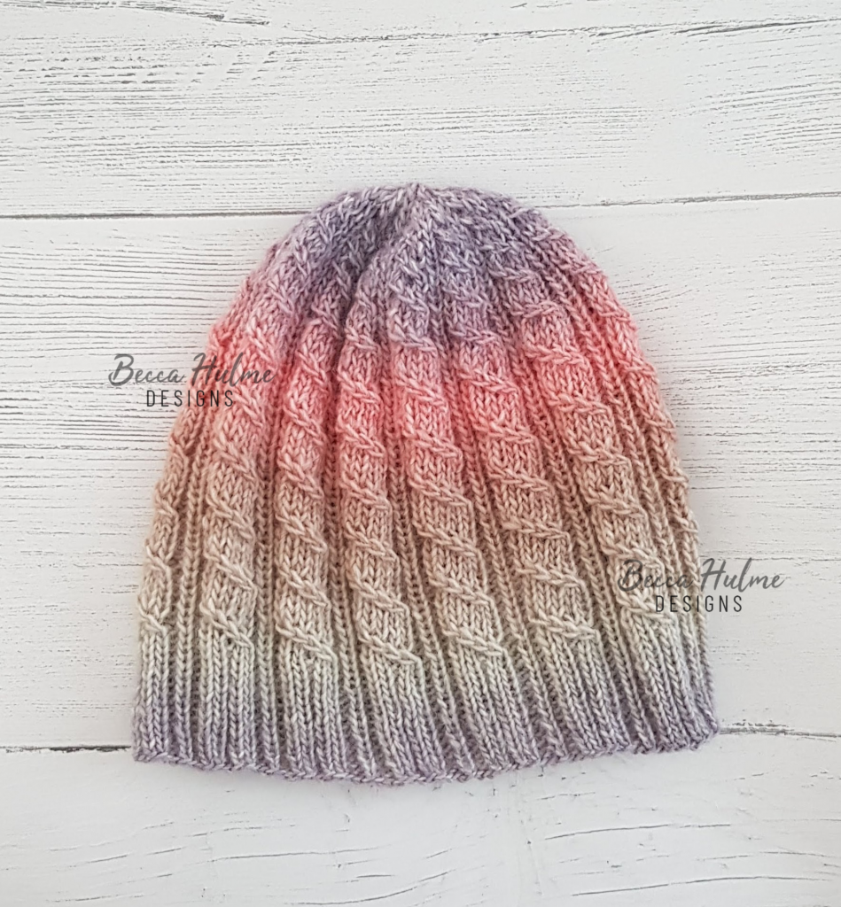 Beanie-style hat laid flat. Colour is a gradient, from light purple to beige, to pink and back to light purple on the crown.