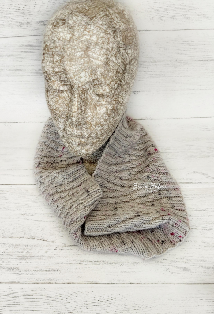 A grey knitted cowl draped around the neck of a mannequin head. The cowl has a chevron effect pattern on it.