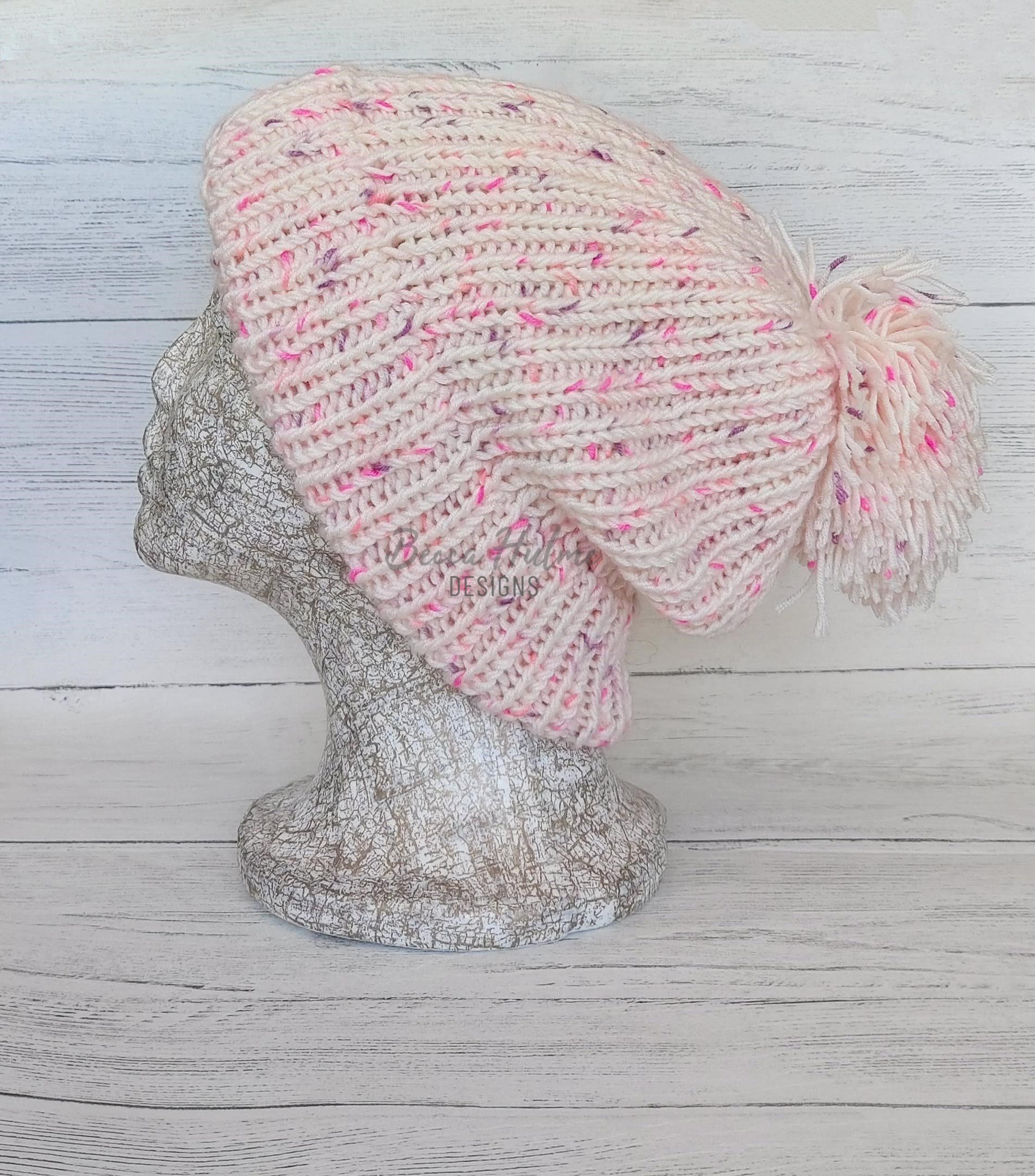 A slouchy beanie-style ribbed knitted hat, pale coloured with pink and purple flecks with a large pompom displayed on a mannequin head.