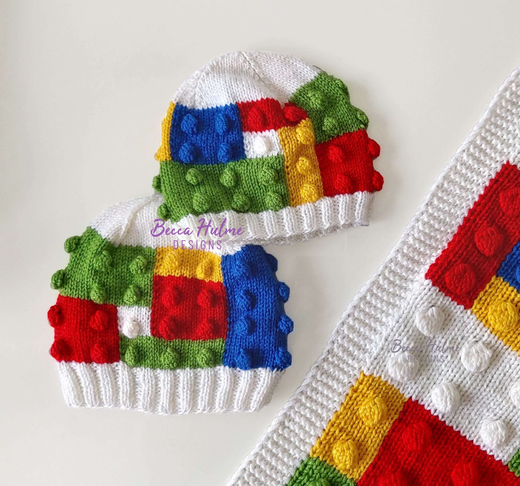 Two colourful knitted hats laid on a table. The design mimics children's building blocks with bobbles and colour layout. Bottom right corner is a part of the Building Blocks Blanket.